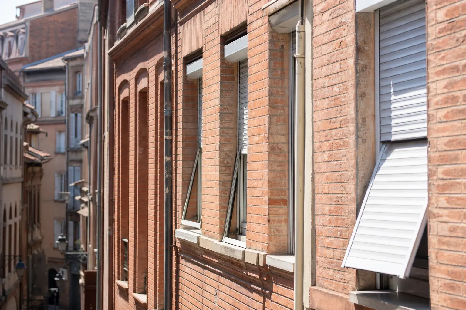 Façade toulousaine en brique rose ancienne avec plusieurs volets roulants aluminium, certains ouverts certains mi-fermés