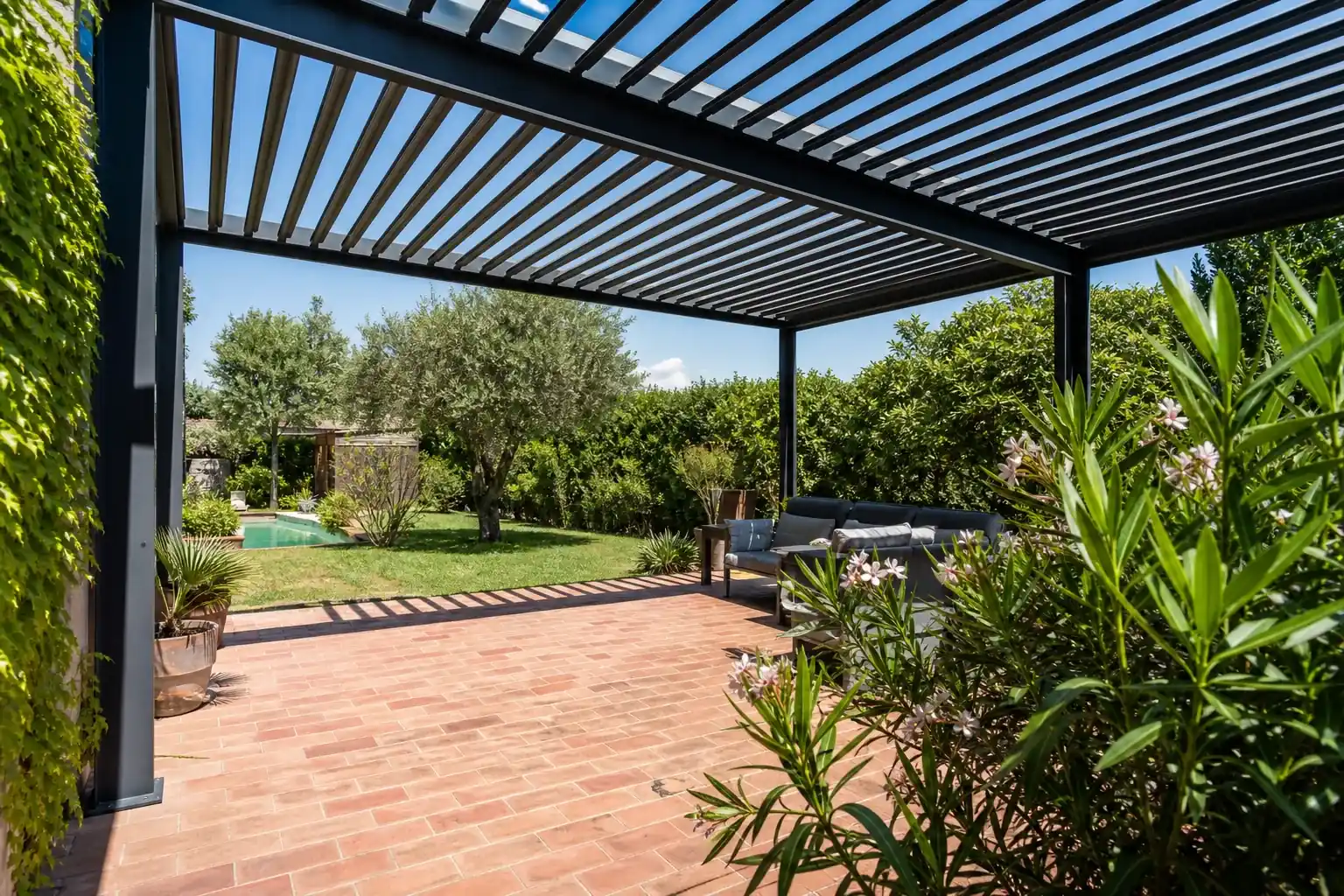 Terrasse pavée de briques roses sous une pergola bioclimatique aux lames grandes ouvertes, plein jour